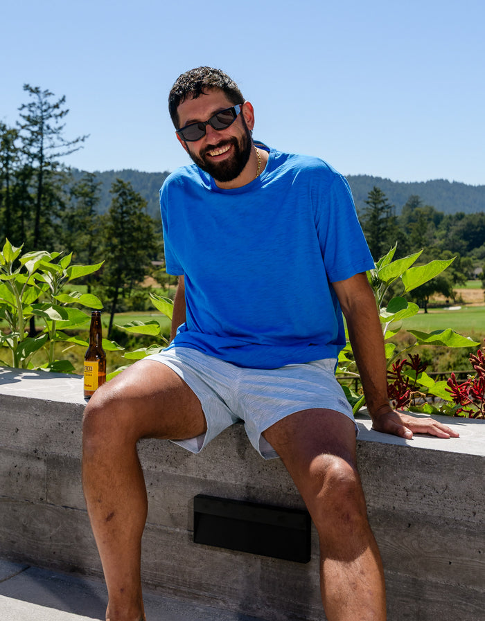 Lifestyle portrait of a man sitting outdoors in a sunny setting, wearing the Baja Llama Solid Blue Slub Pima Tee, showcasing its vibrant color and textured fabric