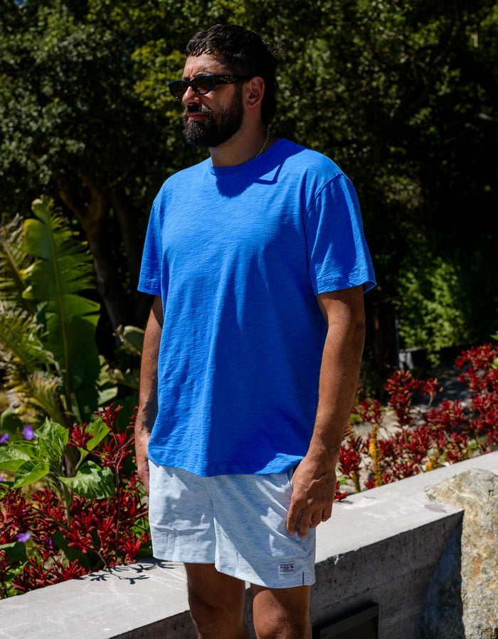A man wearing the Baja Llama Solid Blue Slub Pima Tee, standing in a lush, sunny outdoor garden setting.