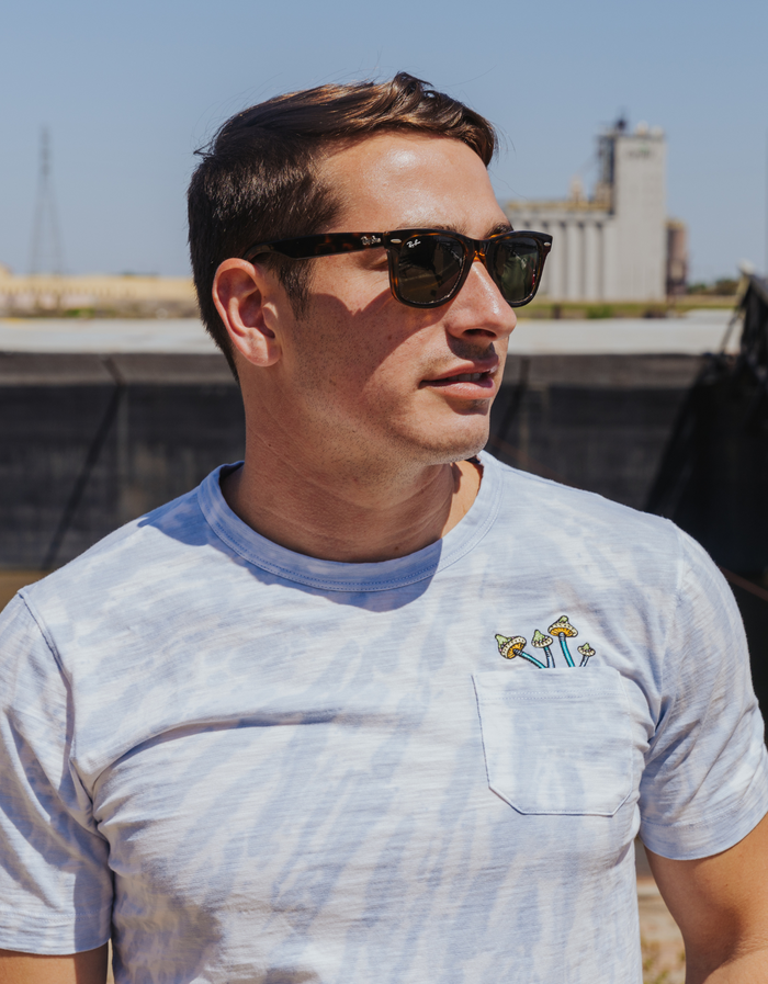 A model wearing the Baja Llama Trip of a Lifetime Slub Pima Pocket Tee, featuring a blue tie-dye design and mushroom embroidery on the pocket, shot in an outdoor lifestyle setting.