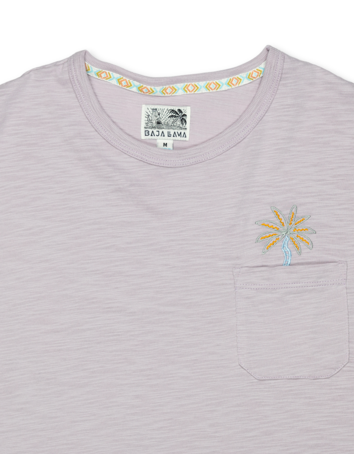 A close-up shot of the neckline and chest on the Baja Llama Marrakesh Palms Purple Rain Pima Slub Pocket Tee, showing the textured lavender purple fabric, a stitched chest pocket with embroidered palm tree, and a geometric pattern lining the inner collar.
