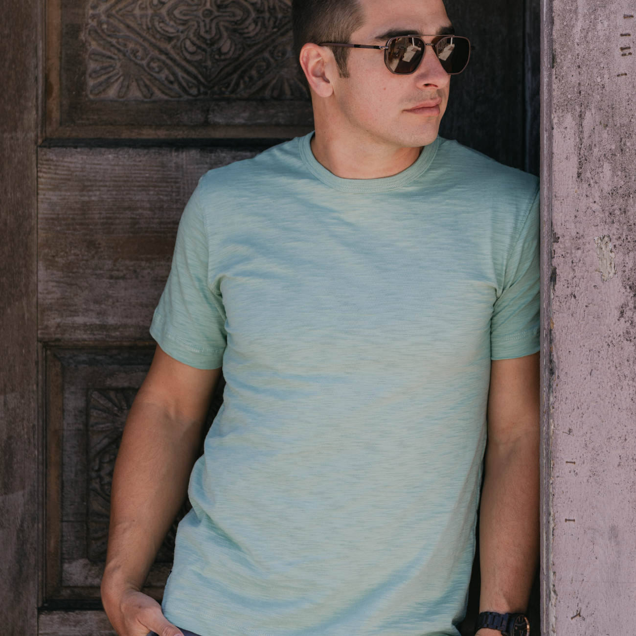 Man wearing Baja Llama Solid Green Slub Pima Pocket Tee – Sage green crewneck T-shirt made from ultra-premium Peruvian cotton with textured slub finish – Styled casually while standing against rustic wooden door – Eco-friendly, breathable, and stylish casual wear for men