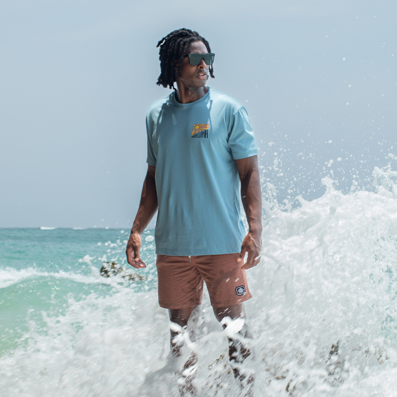 Man wearing Baja Llama Take A Hike Primo Graphic Tee – Teal T-shirt with Stylized Mountain and Sunburst Design – Standing on Coastal Rocks with Ocean Waves Splashing – Premium Peruvian Cotton Adventure Shirt for Men – Eco-Friendly, Ultra-Soft Hiking and Outdoor Lifestyle Apparel