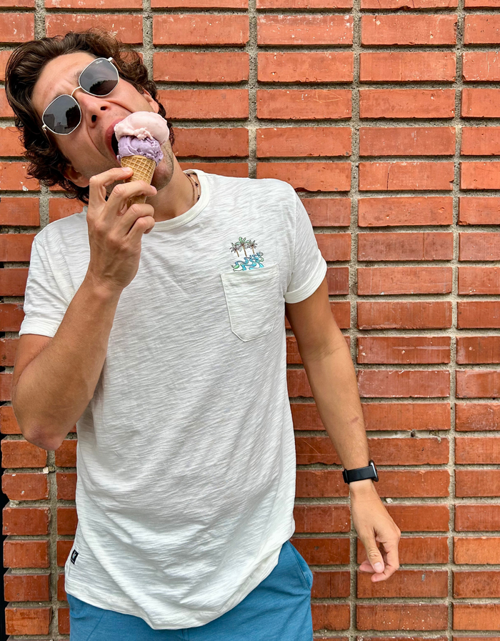 Man wearing the white Baja Llama Palmy Waves Slub Pima Pocket Tee while eating ice cream in front of a brick wall