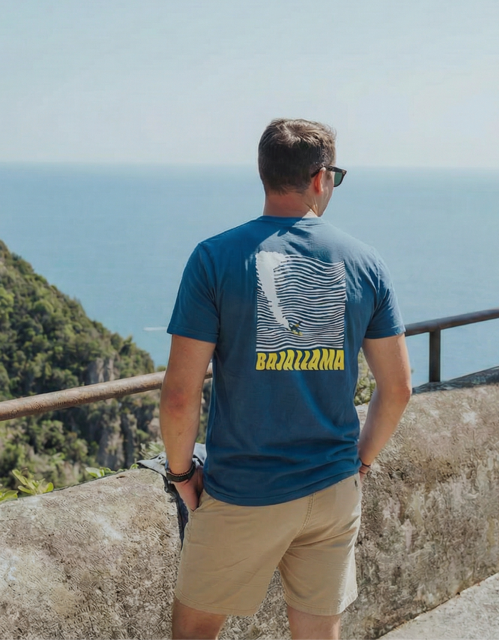 Back view of a man wearing the blue Baja Llama 100 Foot Wave Primo Graphic Tee while overlooking the ocean.