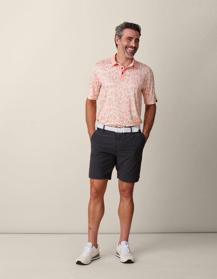 A man smiling with hands in his pockets, wearing the tucked-in Baja Llama A Swarm of Erratic Animals Orange Clay Swingers Performance Polo with a white belt, charcoal gray shorts, and cream sneakers.