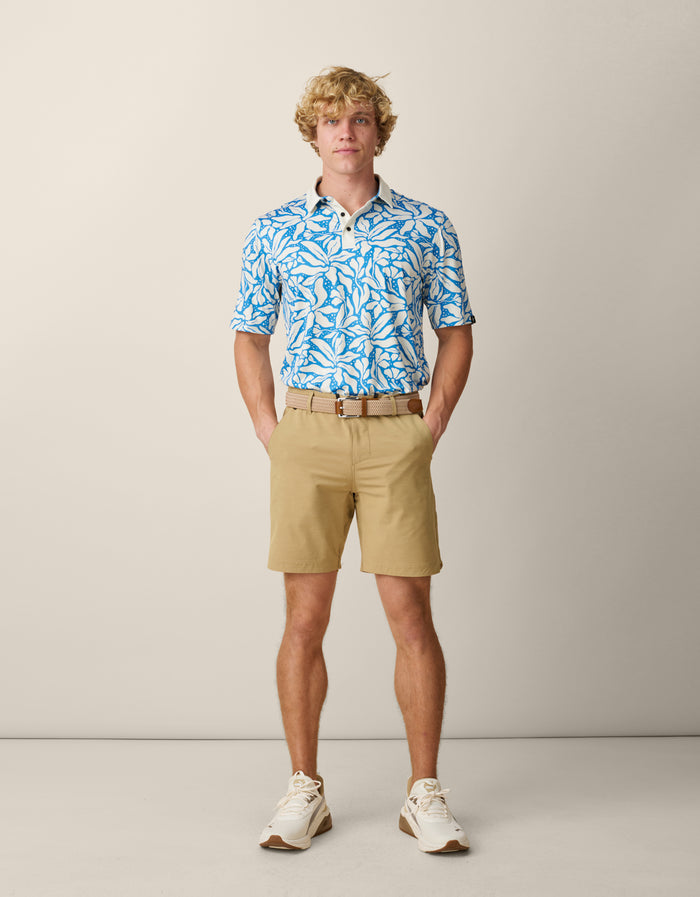 A blonde male model standing with his hands in his pockets, wearing the tucked-in Baja Llama Leaf Me Blue Swingers Performance Polo paired with a beige woven belt, khaki shorts, and cream sneakers.