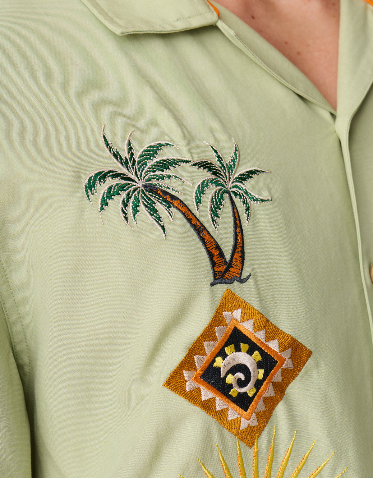 A close-up view of the sage green Baja Llama 'Oasis Geometry' One-Off button-up shirt, showcasing intricate embroidery of palm trees above a geometric diamond-shaped pattern.