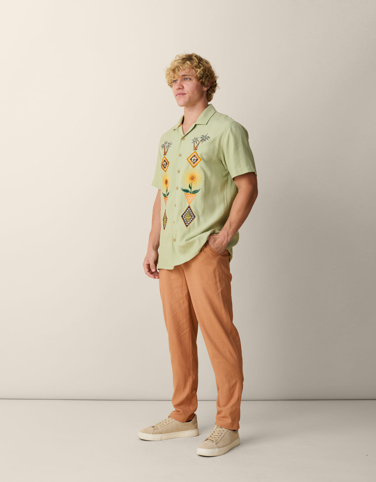 A side-profile view of a male model standing against a neutral studio backdrop, wearing the sage green Baja Llama 'Oasis Geometry' One-Off button-up shirt featuring symmetrical, colorful embroidery, paired with tan trousers and light-colored sneakers.
