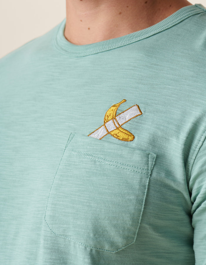 A close-up shot of the left chest on the Baja Llama La Banana Embroidered Pima Slub Pocket Tee as worn by a model, showing the textured sage green fabric topped with a playful embroidered graphic of a banana fixed with white duct tape topped with a chest pocket