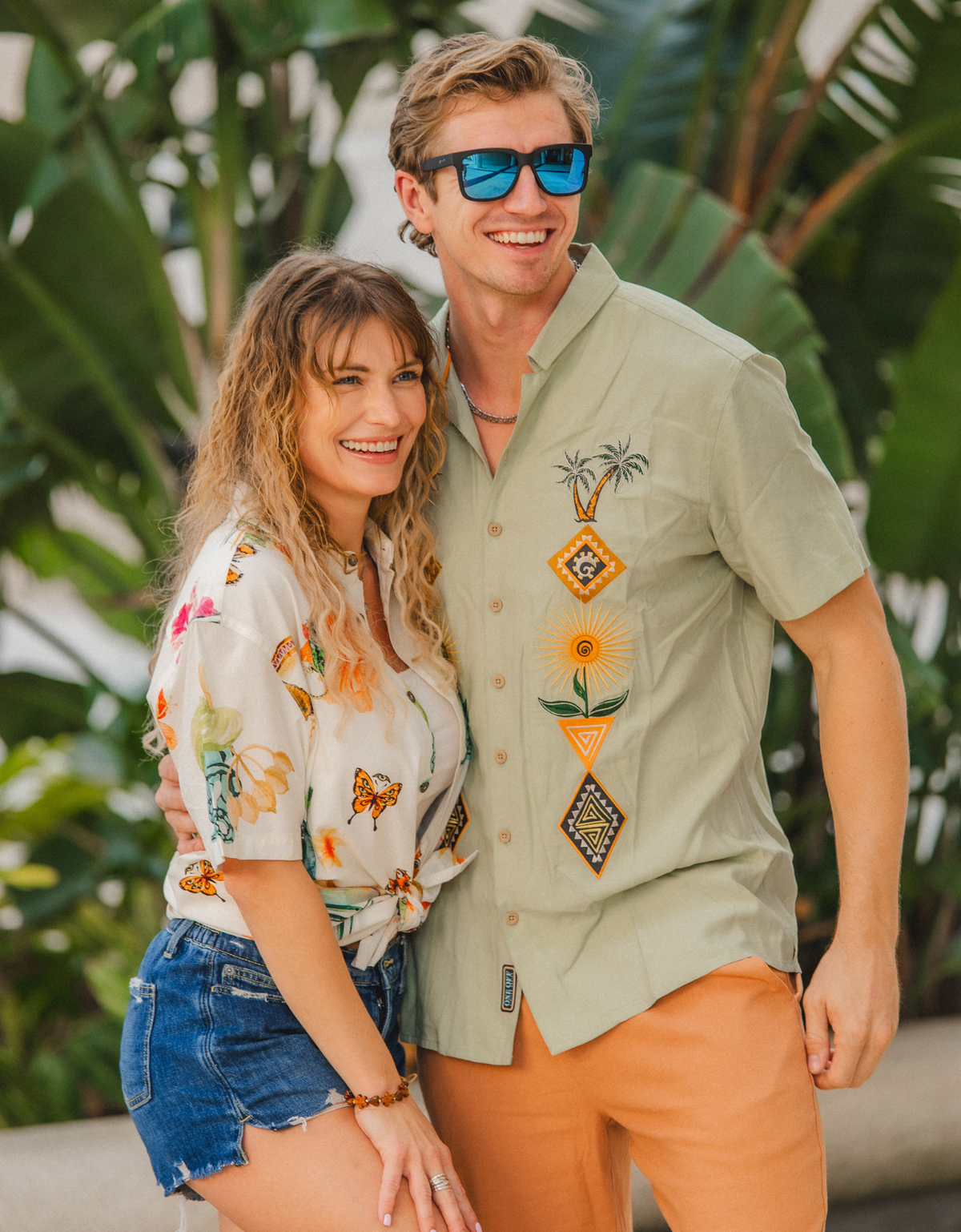 Lifestyle shot of a man wearing the sage green Baja Llama 'Oasis Geometry' One-Off button-up shirt with colorful embroidered desert motifs, standing with a woman in a tropical setting.