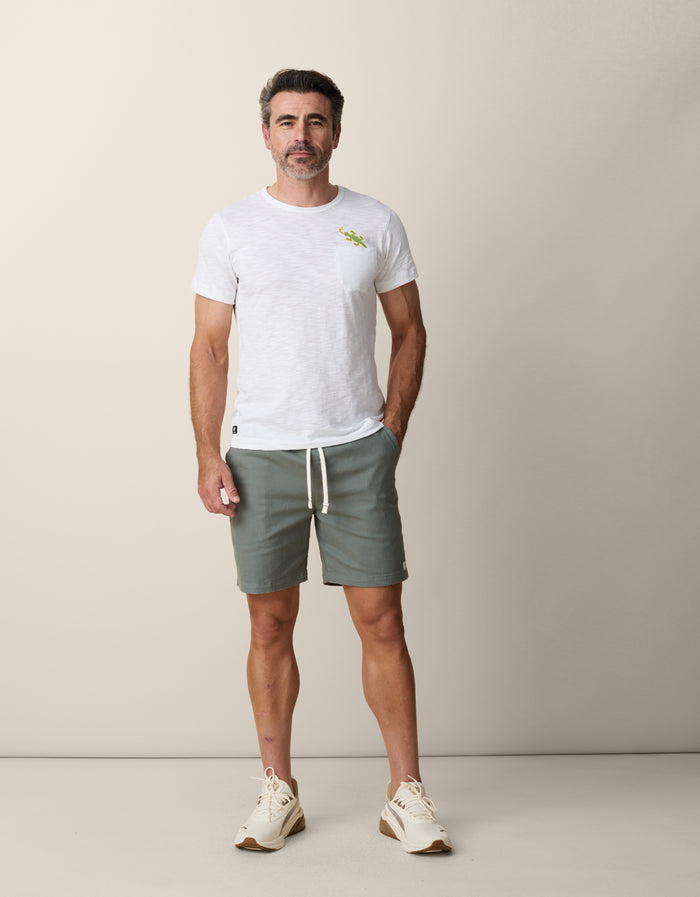 A male model with salt-and-pepper hair standing and facing forward with one hand in his pocket, wearing the untucked Baja Llama Thin Lizzy Pima Slub Pocket Tee in white paired with sage green drawstring shorts and cream sneakers.