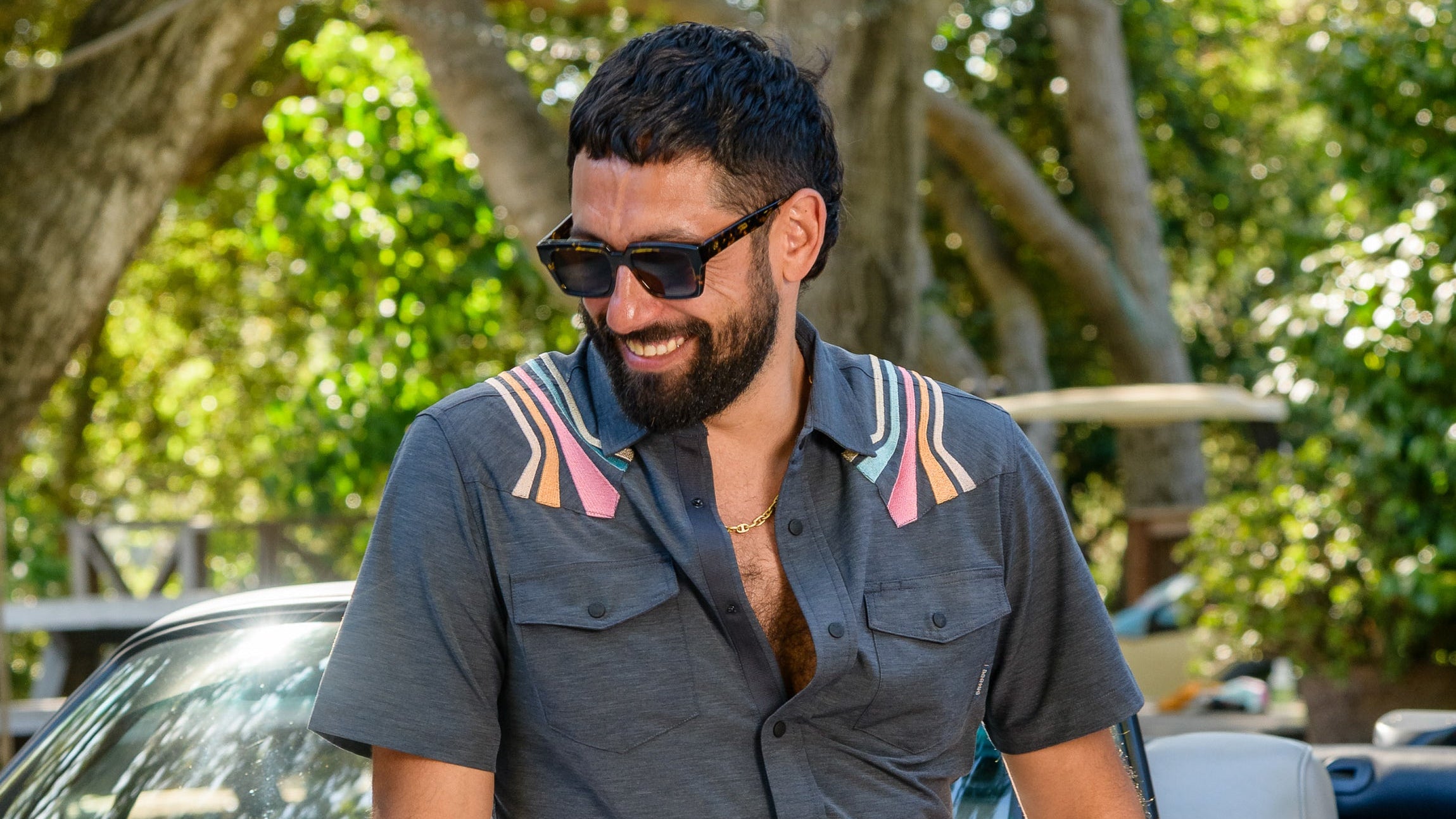 A man smiling and wearing the Baja Llama Retro Rays Black Dissident Western front snap button-up shirt, styled with casual shorts while leaning against a convertible car.