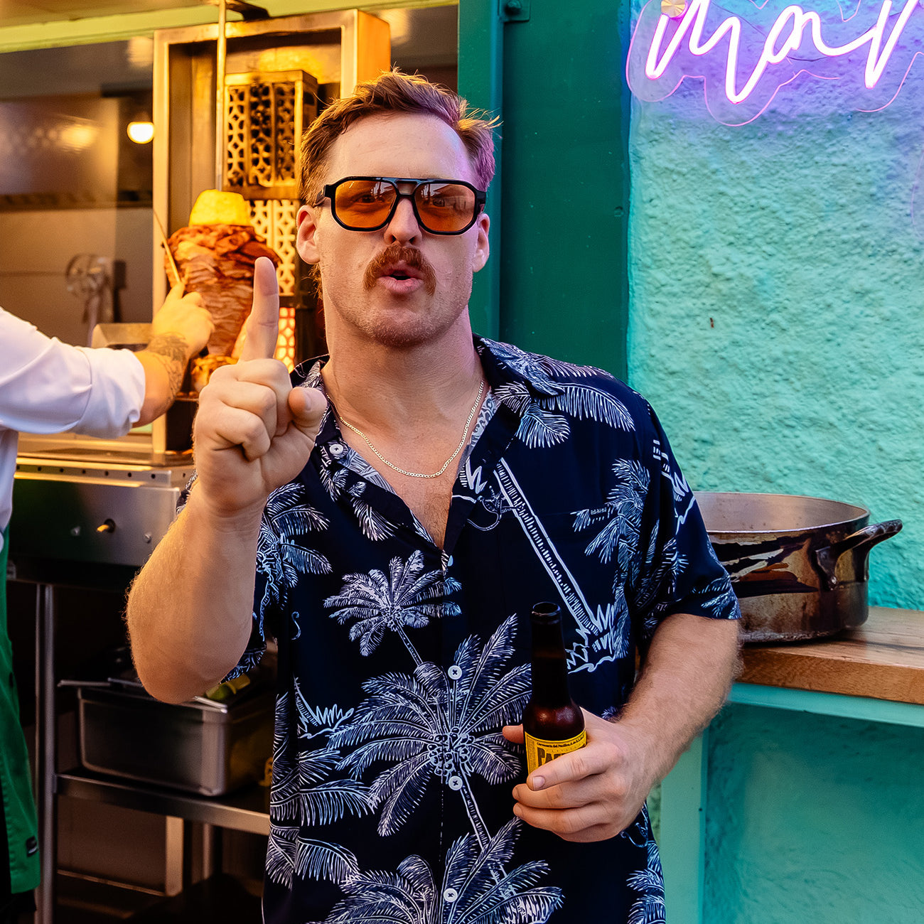 Man wearing Baja Llama The Canopies Are Alive Nighthawk relaxed fit button-up tropical shirt holding a drink in front of a neon sign