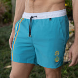 Teal swim shorts with white waistband and a floral and cacti embroidery on a grassy background