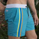 Teal swim shorts with yellow, blue, and orange stripes on a blurred natural background