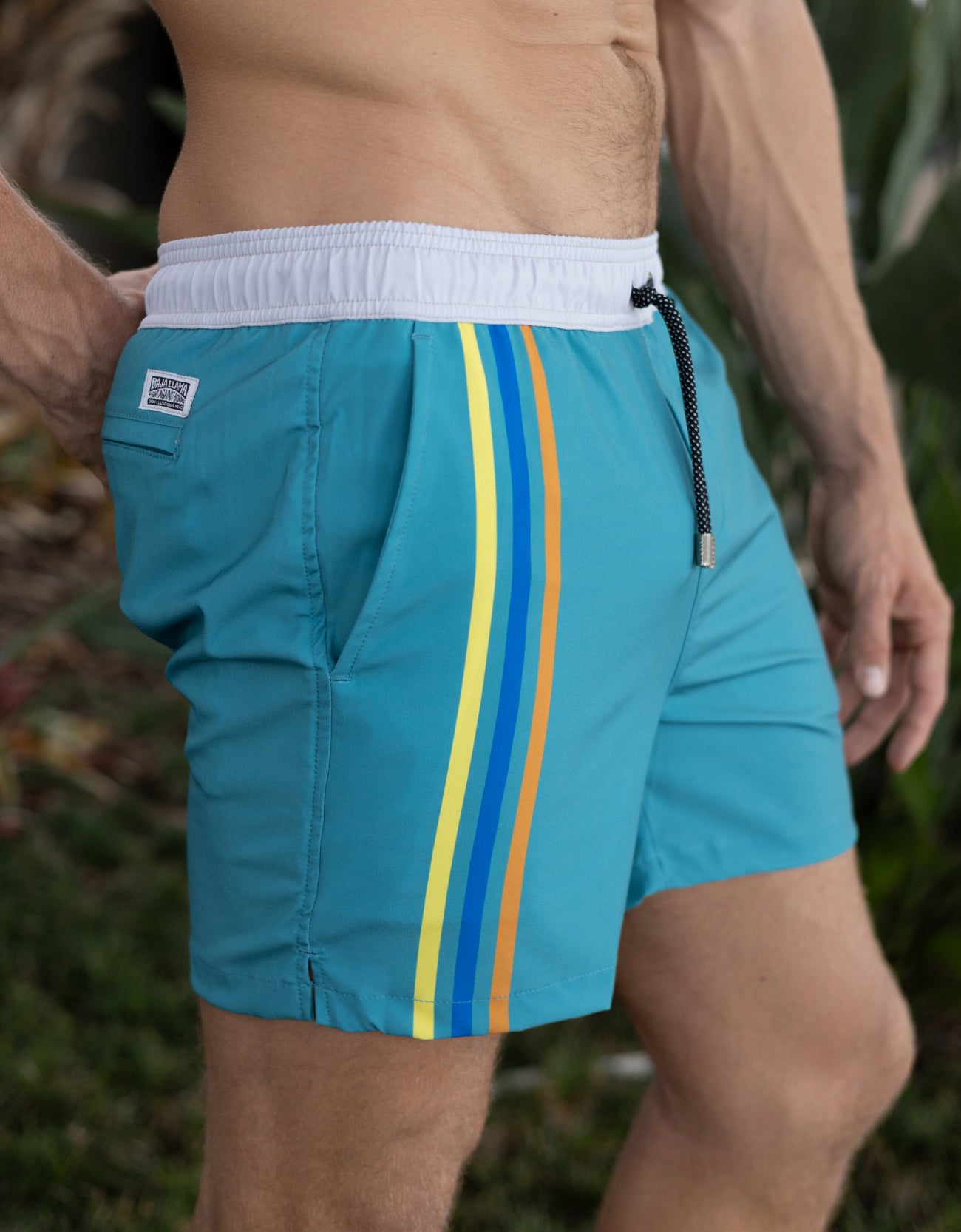 Teal swim shorts with yellow, blue, and orange stripes on a blurred natural background