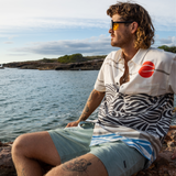 Man wearing Baja Llama Red Sun Over Black Hills Vagabond Button Up shirt by the ocean – artistic modern button-up with abstract waves and red sun design, perfect for travel and coastal adventures