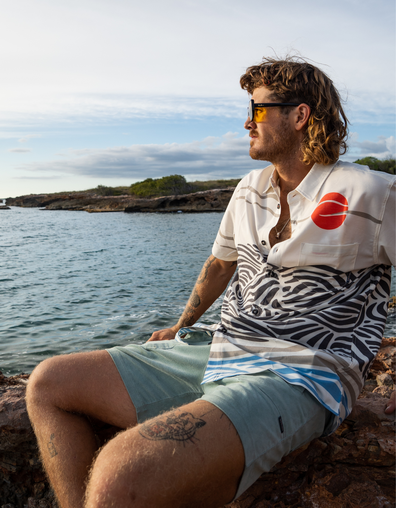 Man wearing Baja Llama Red Sun Over Black Hills Vagabond Button Up shirt by the ocean – artistic modern button-up with abstract waves and red sun design, perfect for travel and coastal adventures