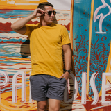 Man wearing Baja Llama Solid Mustard Slub Pima Tee – Mustard yellow crewneck T-shirt made from ultra-soft Peruvian cotton with textured slub finish – Standing against vibrant mural with ‘Paradise’ text – Eco-friendly, breathable, and stylish casual wear for men