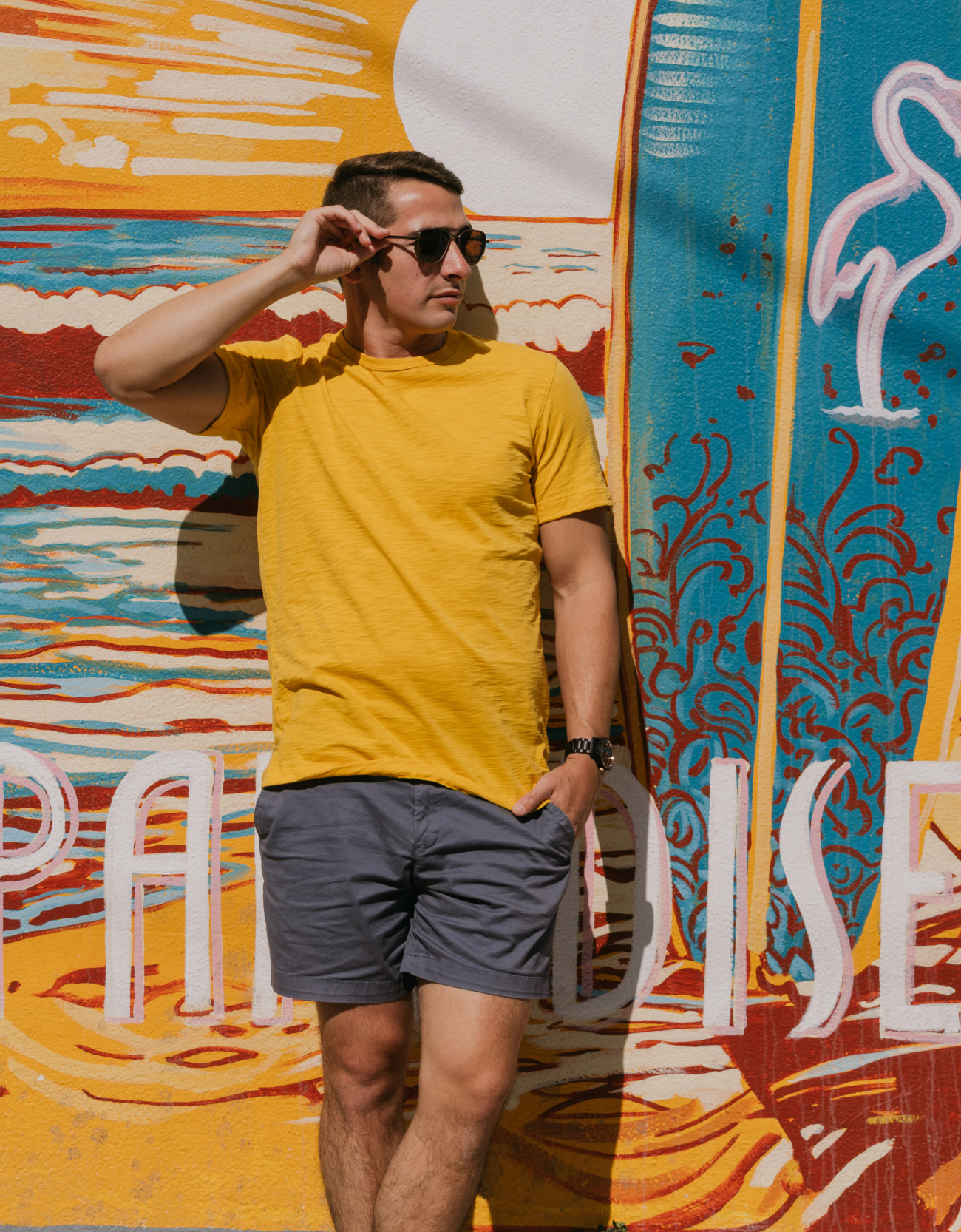 Man wearing Baja Llama Solid Mustard Slub Pima Tee – Mustard yellow crewneck T-shirt made from ultra-soft Peruvian cotton with textured slub finish – Standing against vibrant mural with ‘Paradise’ text – Eco-friendly, breathable, and stylish casual wear for men