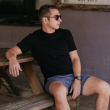Man wearing Baja Llama Solid Black Slub Pima Tee – Black crewneck T-shirt made from ultra-premium Peruvian cotton with textured slub finish – Styled with gray shorts and watch while seated on rustic bench in outdoor setting – Casual, breathable, and eco-friendly men’s fashion for everyday wear