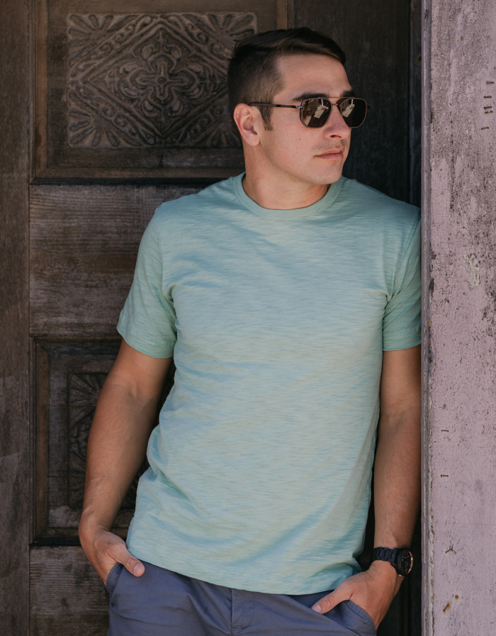 Man by carved door wearing sage Solid Green Slub Pima tee with aviator sunglasses