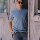 Man wearing Baja Llama Solid Blue Slub Pima Tee – Steel blue crewneck T-shirt made from ultra-premium Peruvian cotton with textured slub finish – Styled with gray shorts while standing against rustic wooden wall  – Eco-friendly, breathable, and stylish casual wear for men