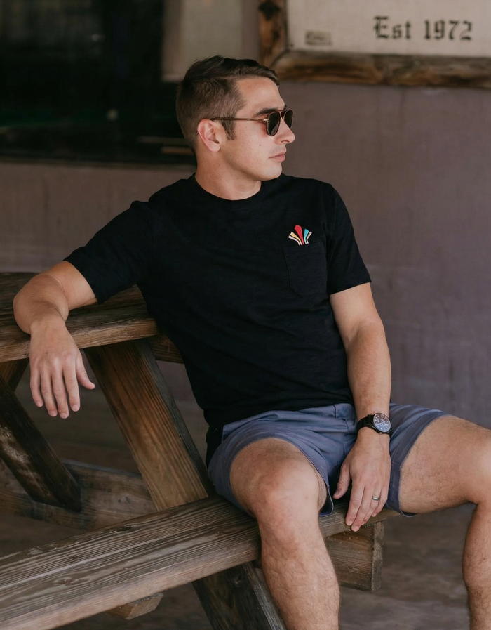 Man wearing a black pocket embroidered t-shirt with a retro logo, sitting on a wooden bench.