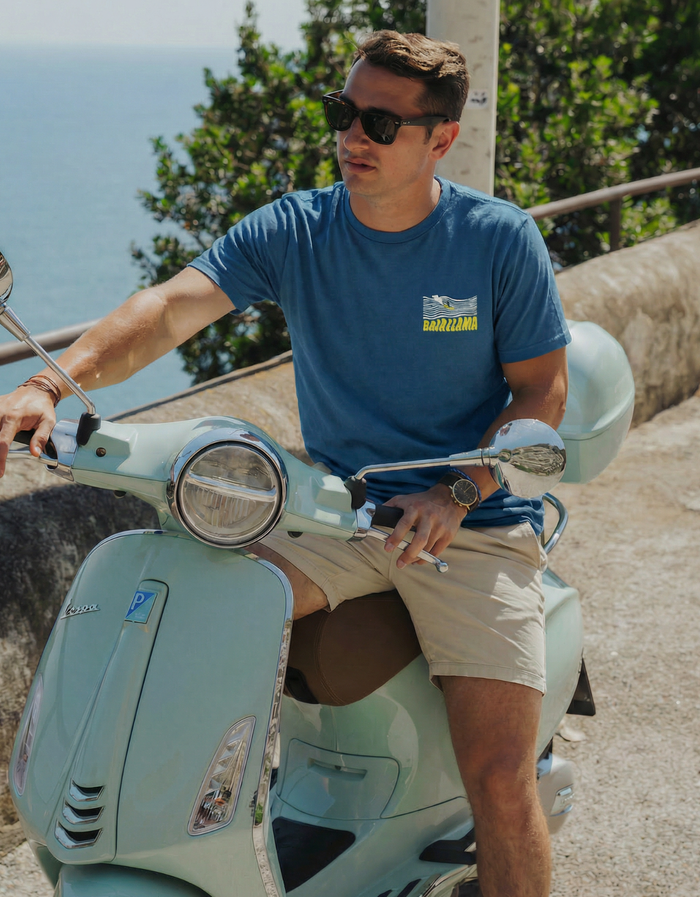 A man wearing the blue Baja Llama 100 Foot Wave Primo Graphic Tee while riding a light blue scooter outdoors.
