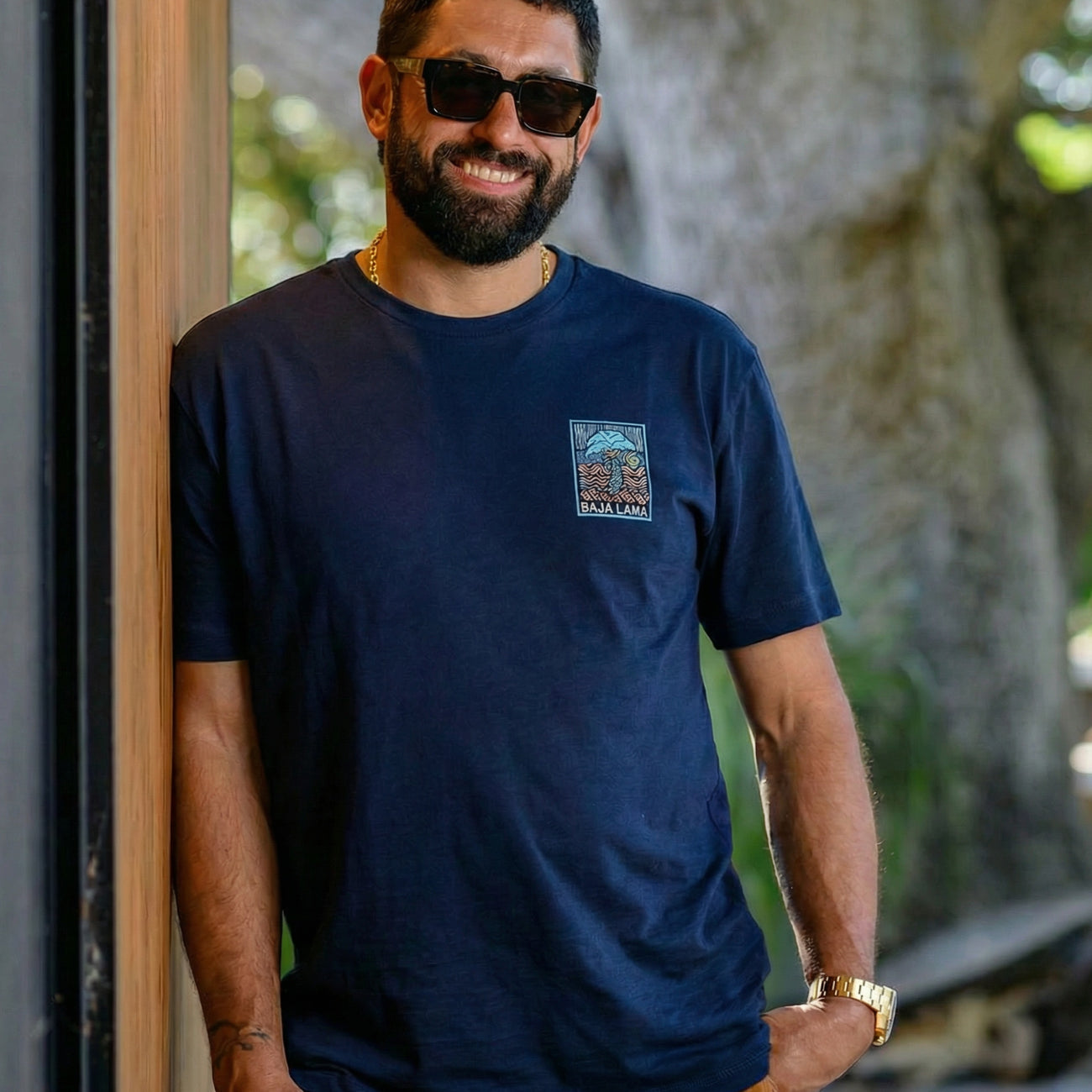 A smiling man with a beard and wearing sunglasses leans  against a wall wearing the Electric Palm - Navy Primo Graphic Shirt