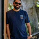 A smiling man with a beard and wearing sunglasses leans  against a wall wearing the Electric Palm - Navy Primo Graphic Shirt