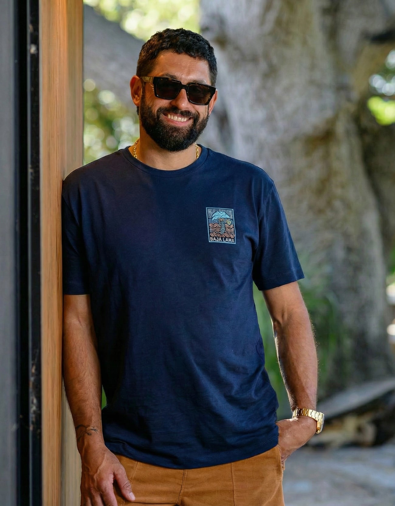 A smiling man with a beard and wearing sunglasses leans  against a wall wearing the Electric Palm - Navy Primo Graphic Shirt
