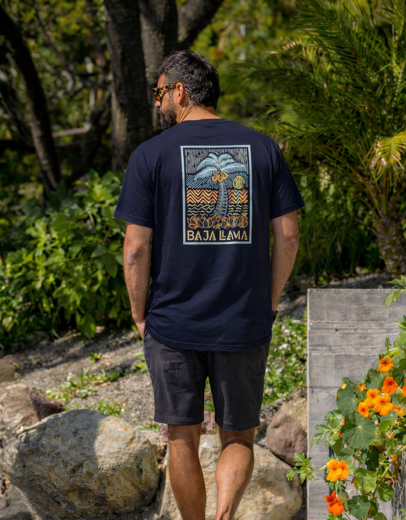 Rear view of a man walking outdoors showcasing the large Electric Palm graphic on the back of his navy blue Baja Llama Primo Graphic Tee