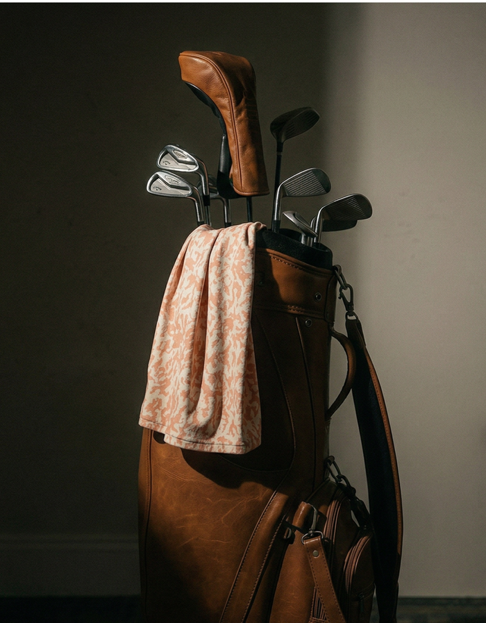 A premium brown leather golf bag filled with clubs, featuring the cream and orange clay patterned Baja Llama A Swarm of Erratic Animals Performance Polo draped neatly over the side.