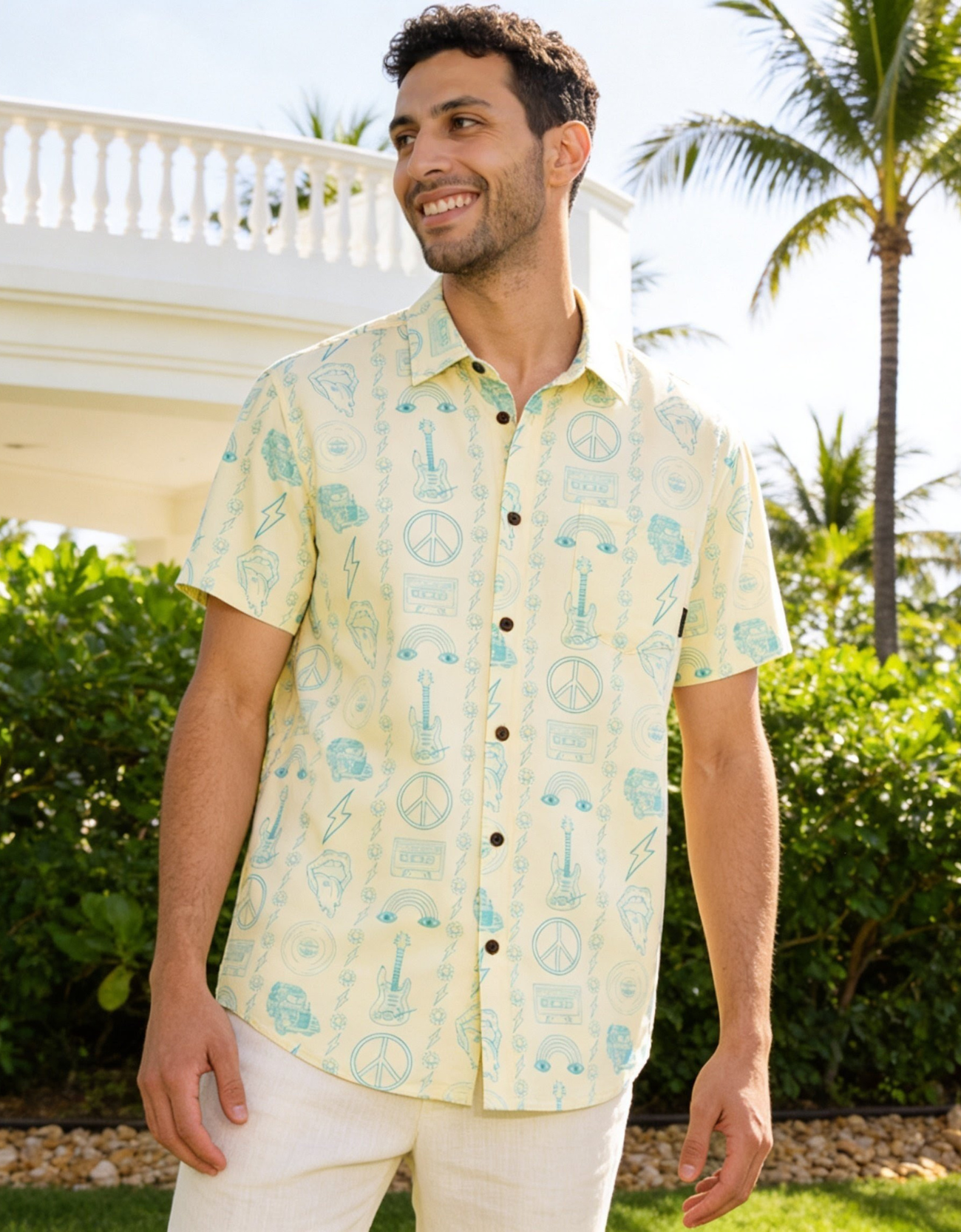 Man wearing a music patterned yellow shirt standing outdoors with greenery and a gazebo in the background
