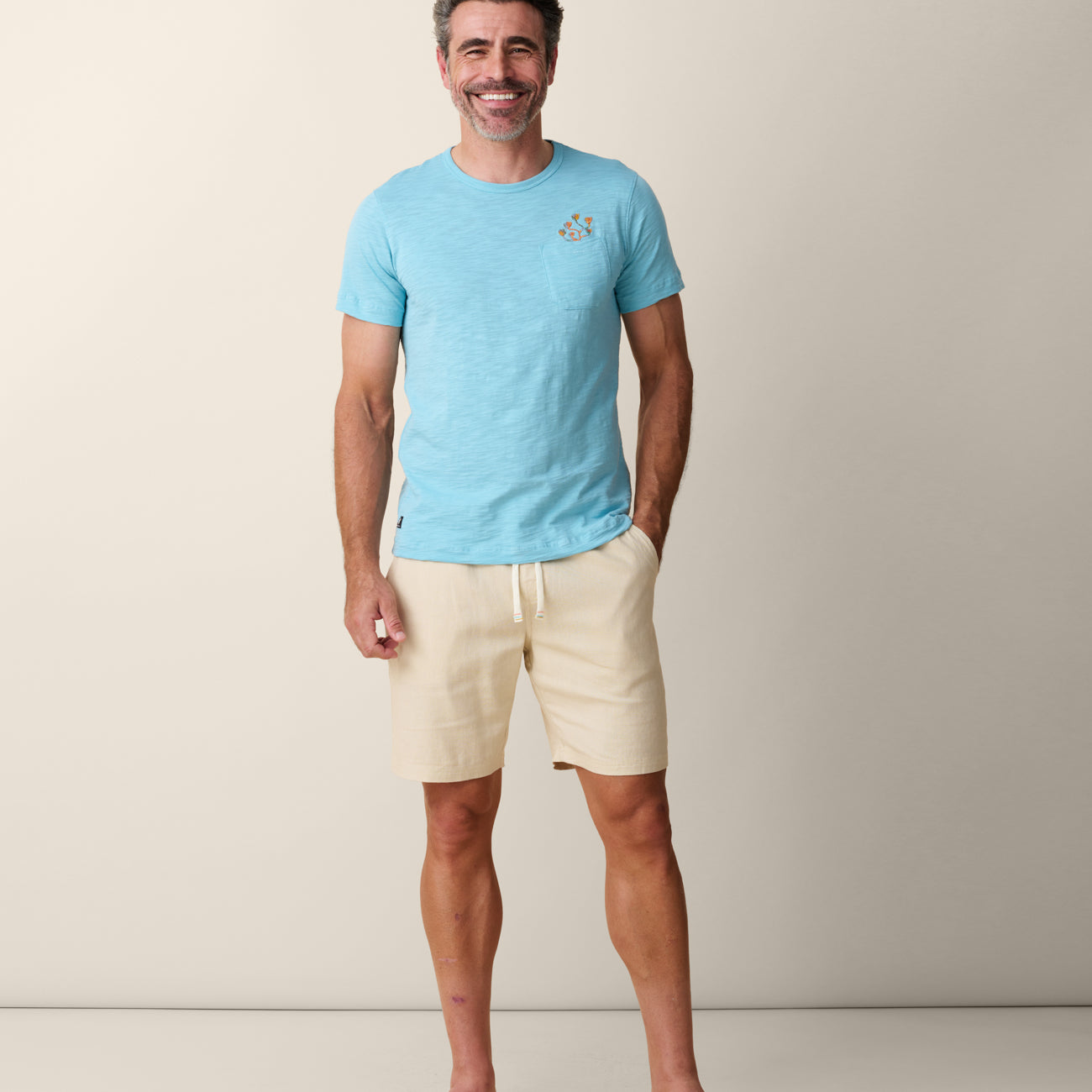 A smiling male model with salt-and-pepper hair standing from the front with one hand in his pocket, wearing the untucked Baja Llama El Sativo Cacti Pima Slub Pocket Tee in porcelain blue, paired with cream-colored drawstring shorts and brown flip-flops.