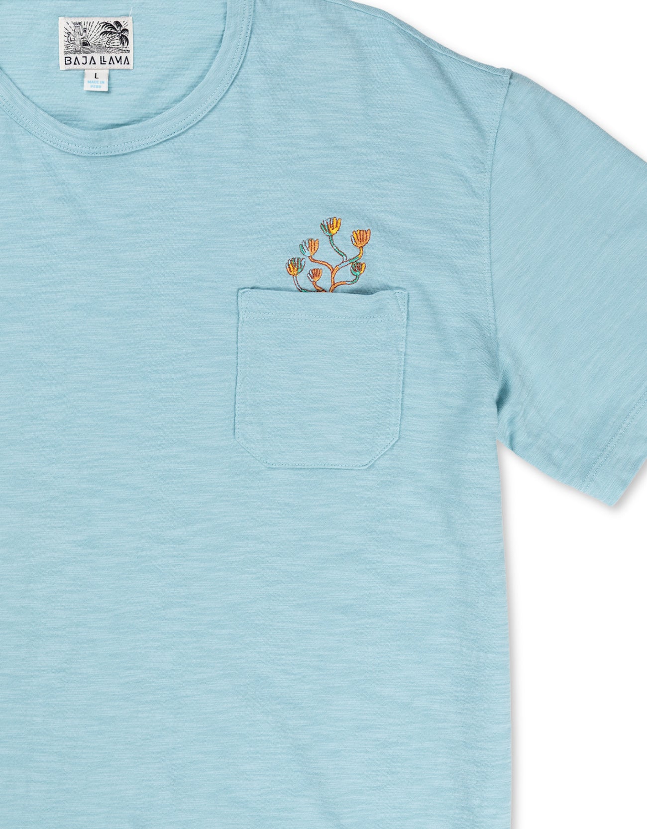 A close-up shot of the neckline and chest area on the Baja Llama El Sativo Cacti Pima Slub Pocket Tee, showing the porcelain blue textured fabric, a patch pocket with an embroidered orange and green cactus
