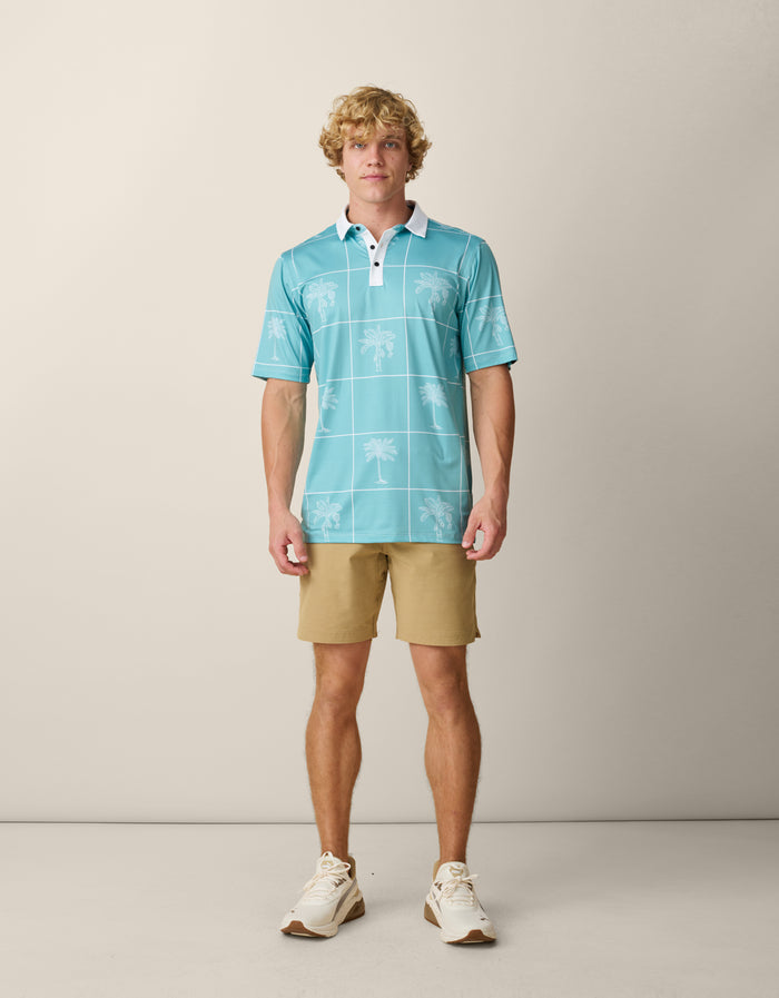 A blonde male model standing and facing forward, wearing the untucked Baja Llama La Republica Piscina Palm Green Swingers Performance Polo paired with khaki shorts and cream sneakers.