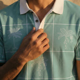 A close-up shot of a man adjusting the collar of the Baja Llama La Republica Piscina Palm Green Swingers Performance Polo, showing the white grid pattern, sketched palm trees, and solid white collar.