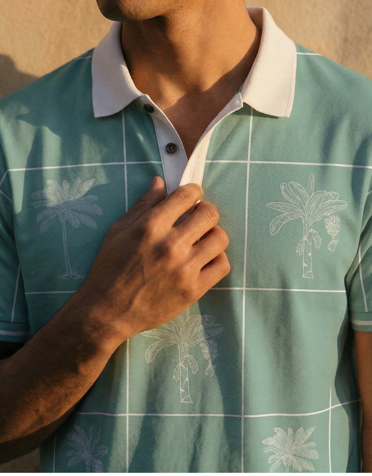 A close-up shot of a man adjusting the collar of the Baja Llama La Republica Piscina Palm Green Swingers Performance Polo, showing the white grid pattern, sketched palm trees, and solid white collar.