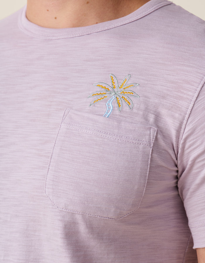 A close-up shot of the left chest on the Baja Llama Marrakesh Palms Purple Rain Pima Slub Pocket Tee as worn by a model, showing the textured lavender purple fabric with an embroidered orange, and blue palm tree topped with a chest pocket.