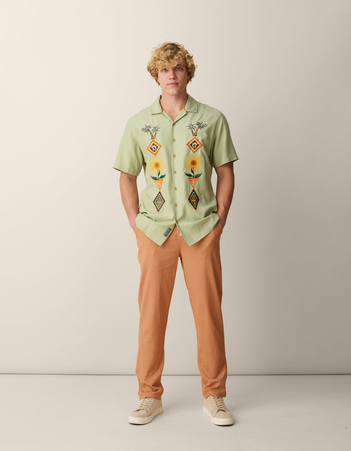 Full-length view of a male model standing against a neutral studio backdrop, wearing the sage green Baja Llama 'Oasis Geometry' One-Off button-up shirt featuring symmetrical colorful desert embroidery, styled with tan trousers and light-colored sneakers.