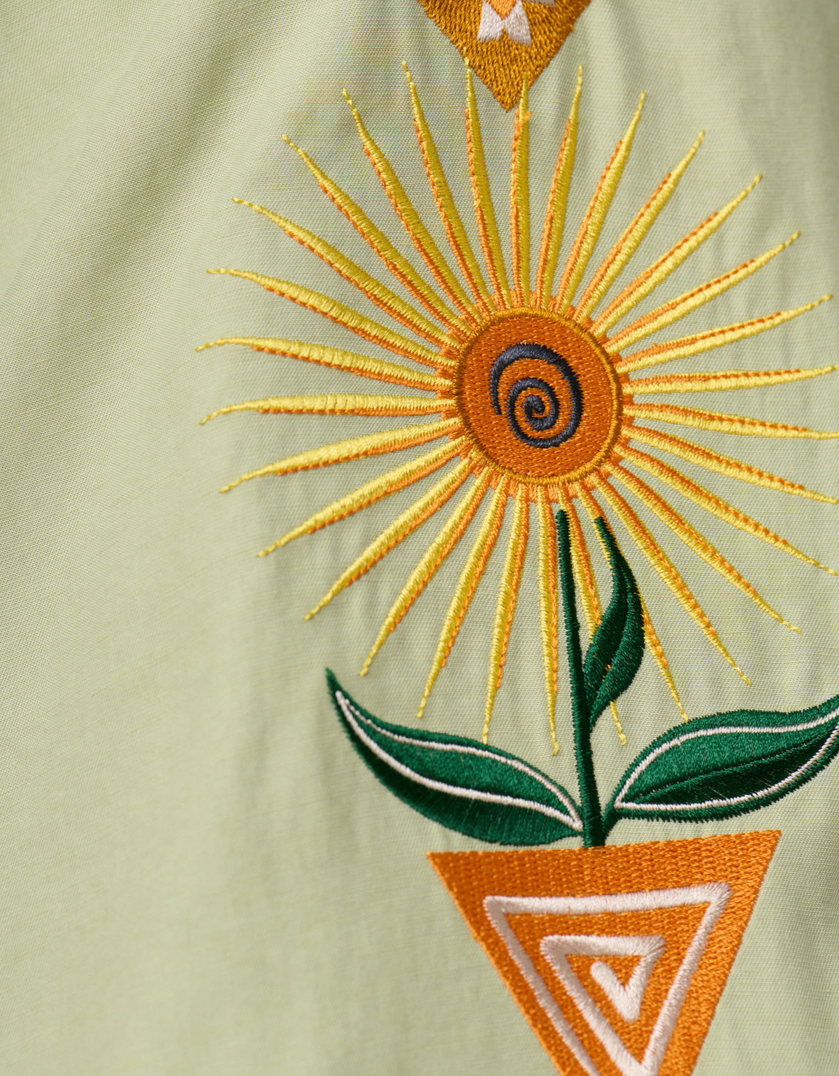 A close-up view of the sage green Baja Llama 'Oasis Geometry' One-Off button-up shirt, highlighting intricate embroidery of a vibrant yellow and orange stylized sun flower growing out of a geometric triangular pot.