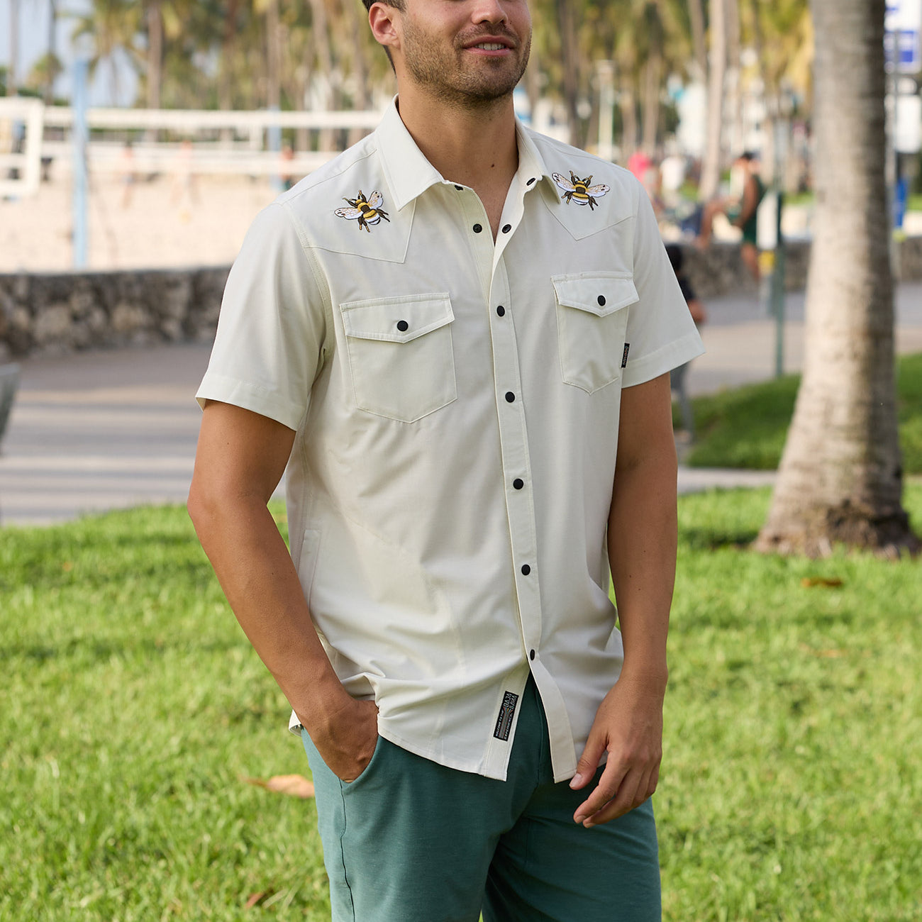 Lifestyle photo of a man wearing a cream-colored short-sleeve western snap shirt with embroidered bee details near a park