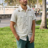 Lifestyle photo of a man wearing a cream-colored short-sleeve western snap shirt with embroidered bee details near a park