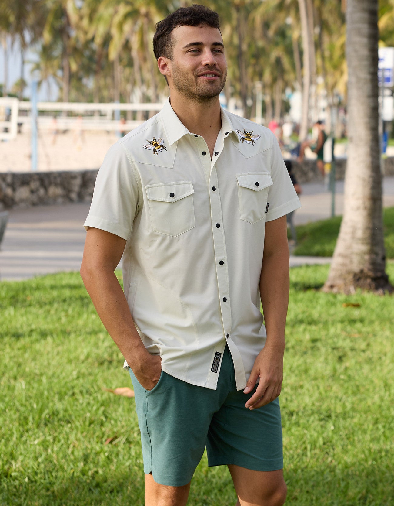 Lifestyle photo of a man wearing a cream-colored short-sleeve western snap shirt with embroidered bee details near a park