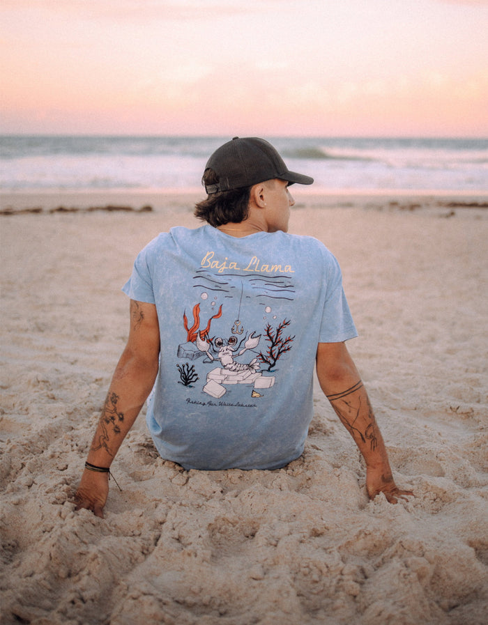 Model sitting on the beach wearing the Baja Llama White Lobster Acid Wash Primo Graphic Tee, showing the large lobster artwork on the back.