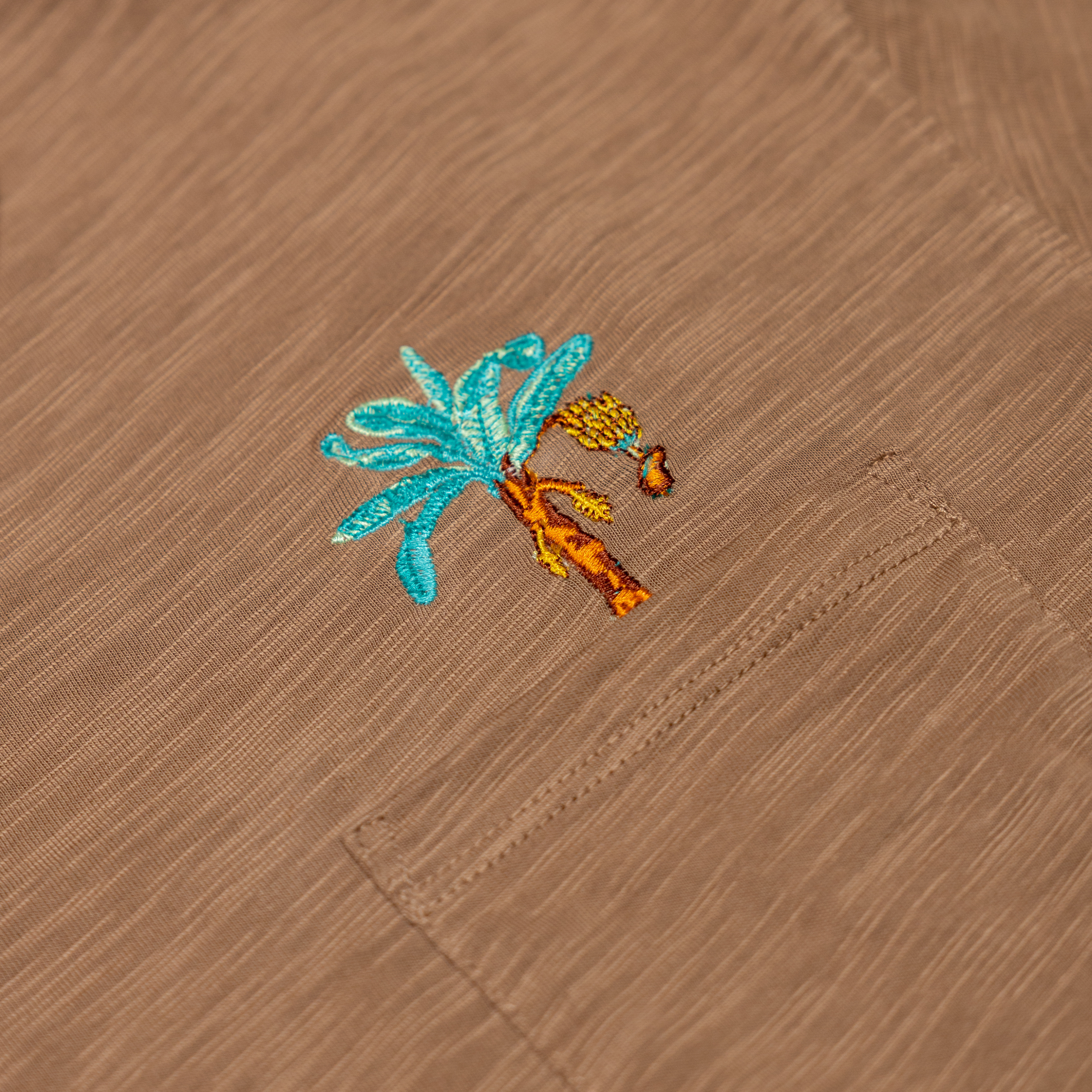 An extreme close-up shot of the left chest pocket area on the Baja Llama La Republica Embroidered Pima Slub Pocket Tee, highlighting the detailed brown slub fabric texture and an embroidered turquoise and orange banana tree with a hanging bunch of bananas.