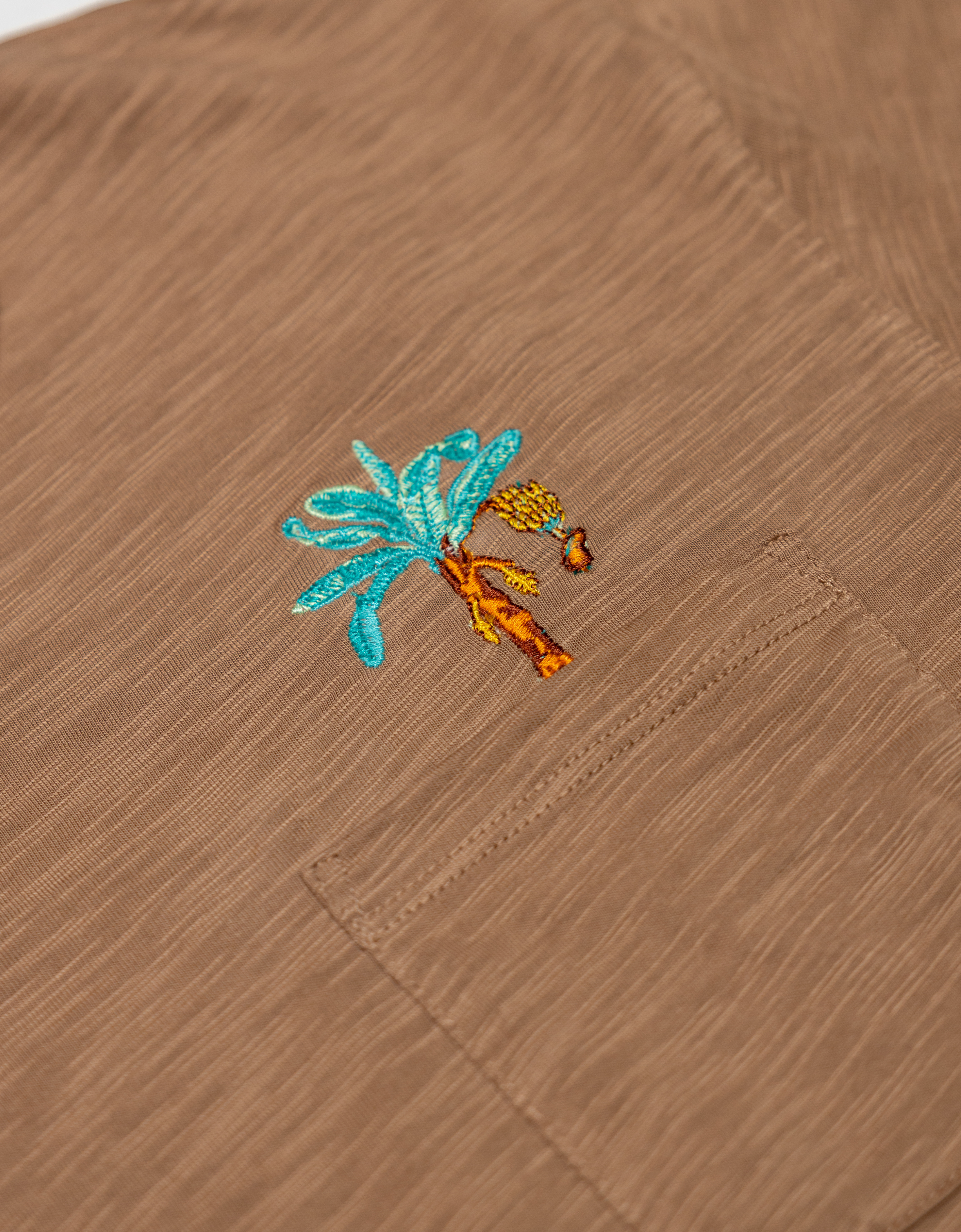 An extreme close-up shot of the left chest pocket area on the Baja Llama La Republica Embroidered Pima Slub Pocket Tee, highlighting the detailed brown slub fabric texture and an embroidered turquoise and orange banana tree with a hanging bunch of bananas.
