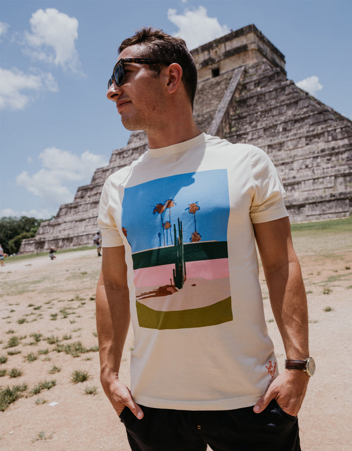 A man wearing the Baja Llama 5PM Primo graphic tee, featuring a retro desert cactus and palm tree landscape print, while standing in front of an ancient pyramid.